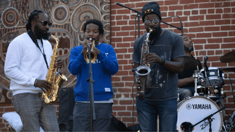 Three men play horns on an outdoor stage