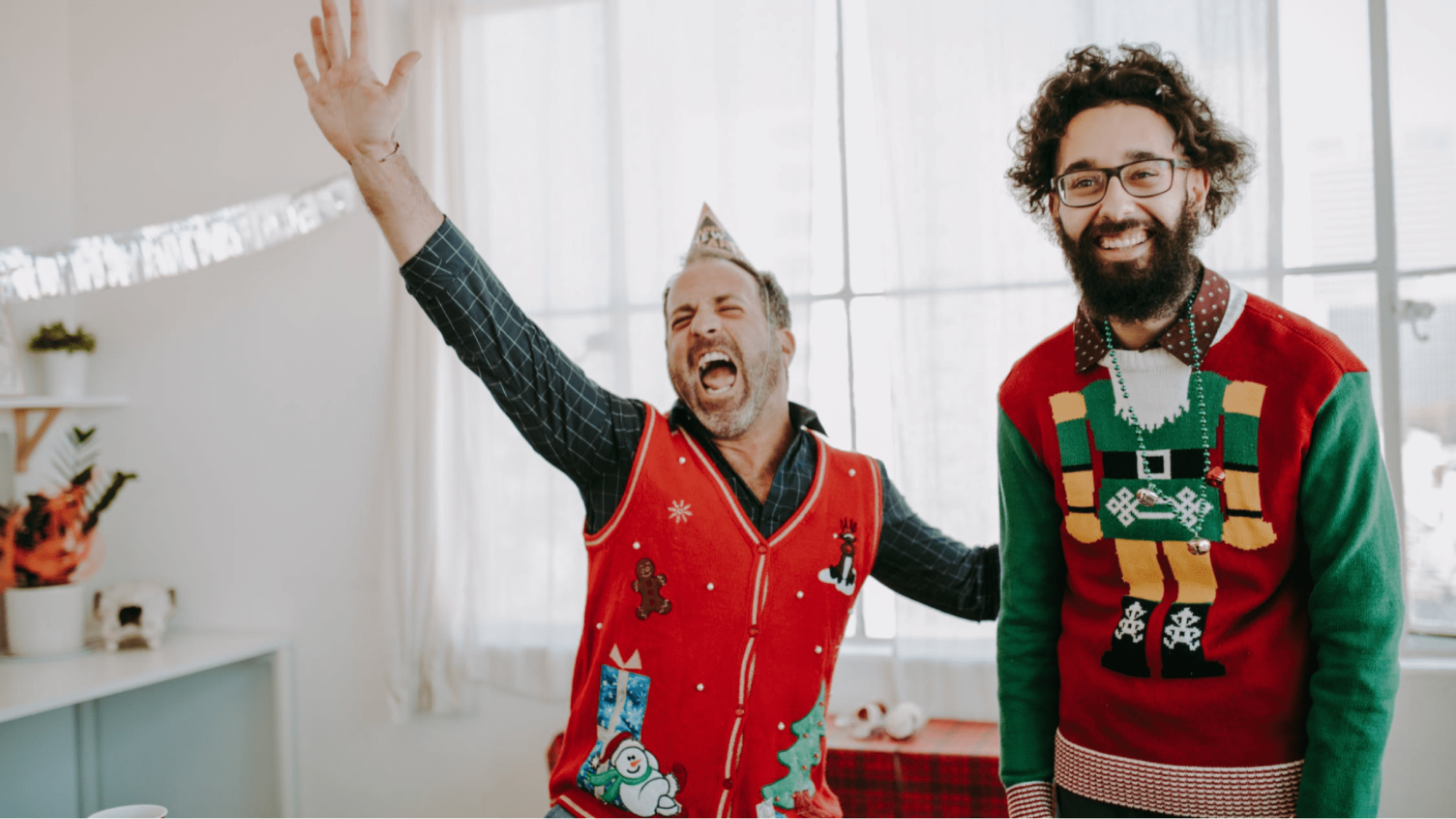 Two men in Christmas sweaters laugh while playing beer pong